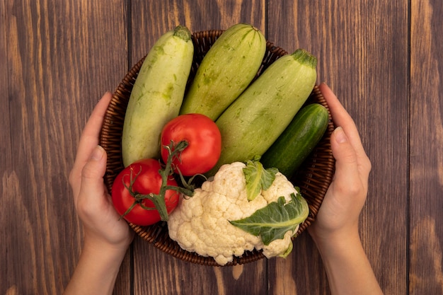 Top view of female hands holding a bucket of fresh vegetables such as zucchinis tomatoes cucumber and cauliflower on a wooden wall