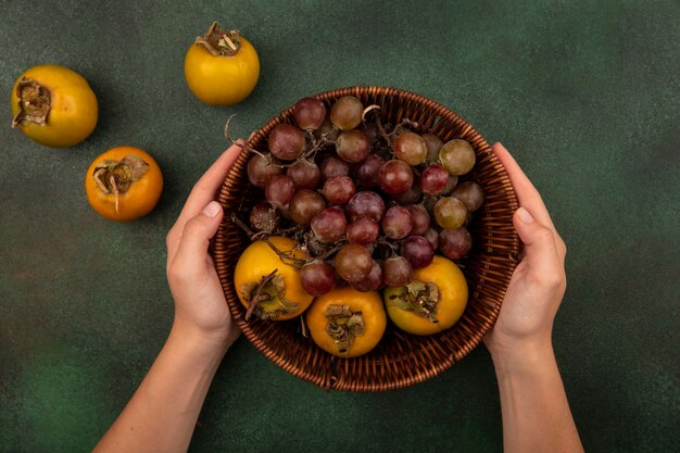 Top view of female hands holding a bucket of persimmon fruits with grapes on a green background