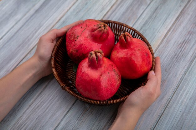 Top view of female hands holding a bucket with red fresh pomegranates on a grey wooden background