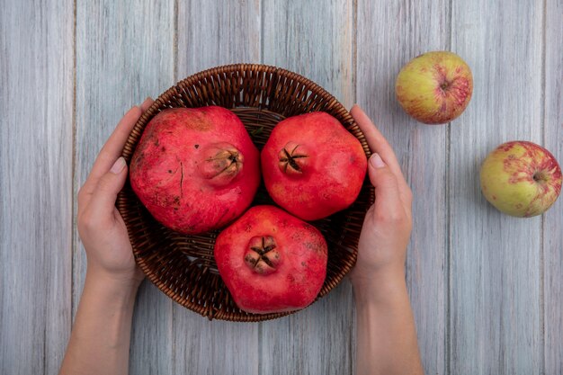 Top view of female hands holding a bucket with red fresh pomegranates with apples isolated on a grey wooden background