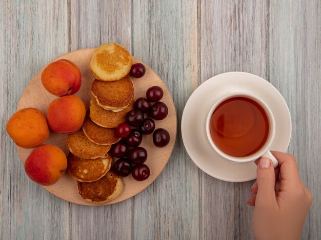 Top view of female hands holding cup of tea and pancakes with apricots and cherries on cutting board on wooden background