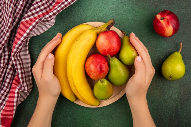 Top view of female hands holding cutting board with fruits on it as peach pear banana with plaid cloth on green background