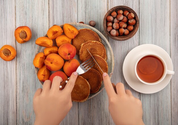 Вид сверху на женские руки, держащие вилку и нож с тарелкой блинов с целыми и нарезанными абрикосами и чашкой чая с миской орехов на деревянном фоне