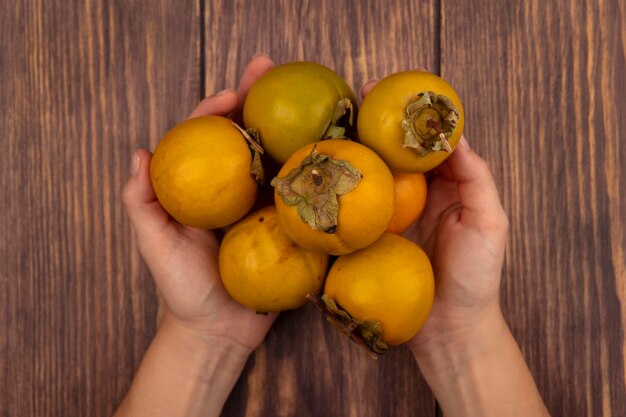 Вид сверху женских рук, держащих свежие оранжевые плоды хурмы на деревянном столе