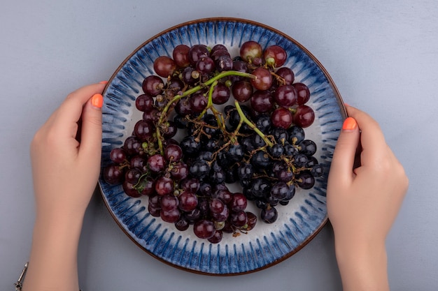 Top view of female hands holding plate of black grape on gray background