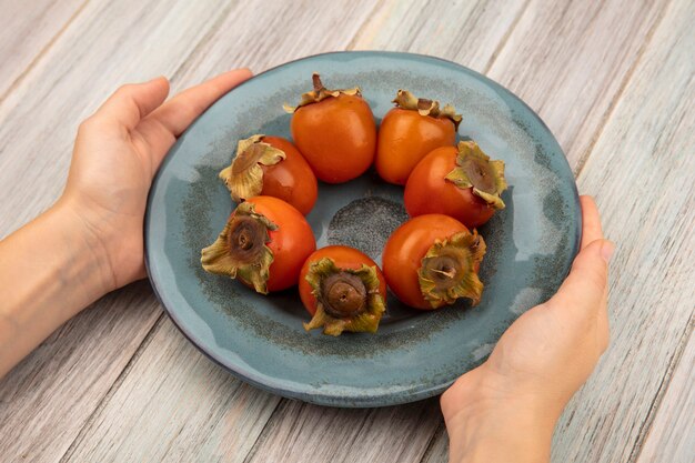 Top view of female hands holding a plate of soft persimmons on a grey wooden surface