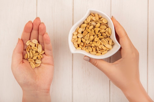 Top view of female hands holding salty pine nuts in one hand and in the other hand the bowl with pine nuts on a beige wooden table