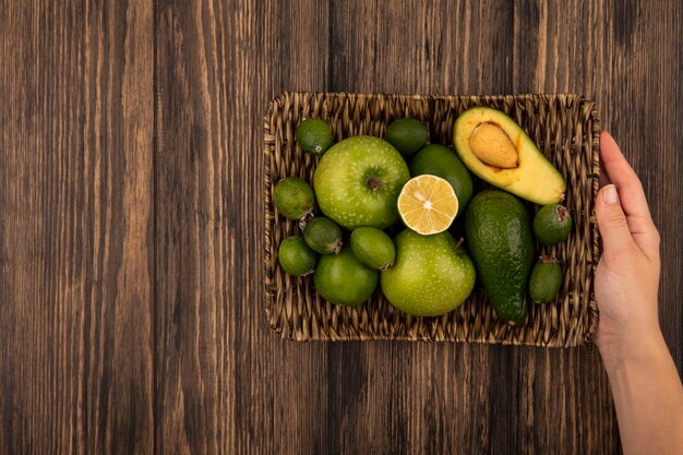 Top view of female hands holding a wicker tray of fresh fruits such as green apples feijoas limes on a wooden wall with copy space