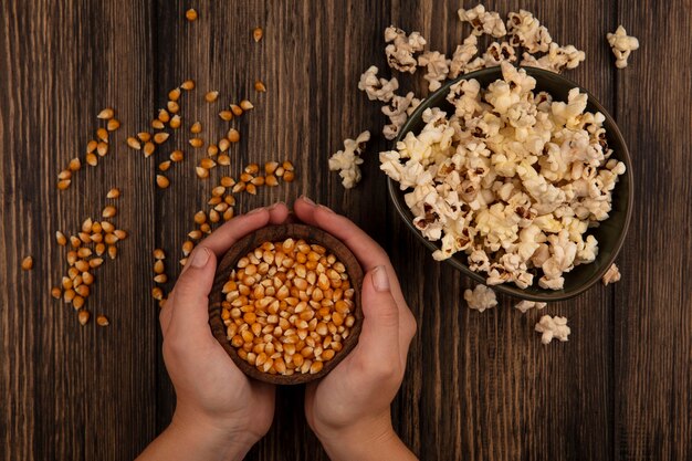 Top view of female hands holding a wooden bowl of corn kernels with popcorns on a bowl on a wooden table
