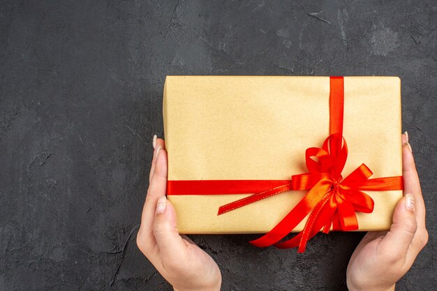 Top view female hands holding xmas gift in brown paper tied with red ribbon on dark surface