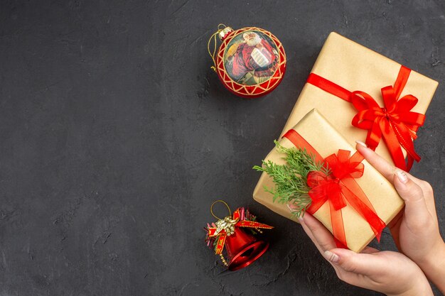 Вид сверху женские руки, держащие рождественский подарок в коричневой бумаге, перевязанные красной лентой, рождественские елочные игрушки на темном фоне, свободное пространство