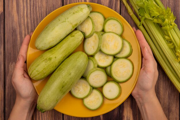 Top view of female hands holding a yellow plate of fresh zucchinis with celery isolated on a wooden surface