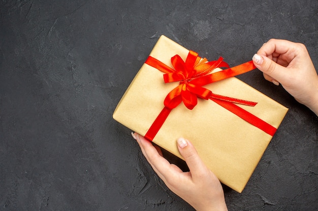 Вид сверху женские руки открывают рождественский подарок в коричневой бумаге с красной лентой на темной поверхности