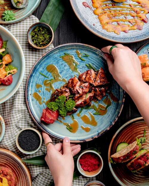 top view of female hands putting a plate with roasted chicken with grilled tomato fresh herbs and sauce on a table