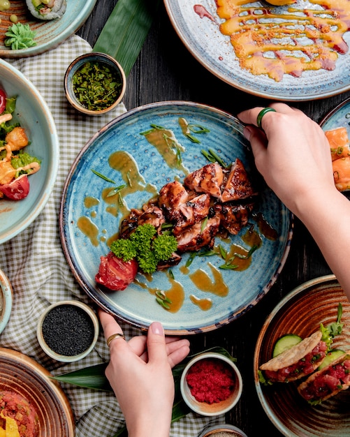 top view of female hands putting a plate with roasted chicken with grilled tomato fresh herbs and sauce on a table