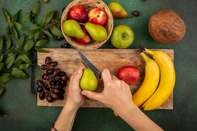 Foto gratuita vista superiore delle mani femminili che affettano la pera con il coltello e l'uva della pesca della banana sul tagliere e la noce di cocco della mela della pera con le foglie su fondo verde