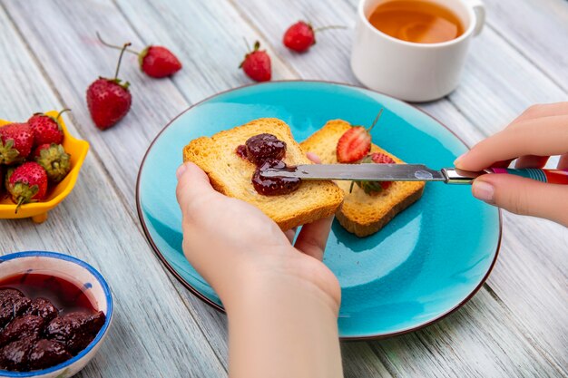 Top view of female hands spread strawberry jam on bread with knife over a blue dish with fresh strawberries on a yellow bowl on a grey wooden background