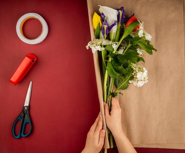 Top view of female hands wrapping a bouquet of blooming viburnum calla lily and dark purple iris flowers with craft paper and scissors, stapler and roll of adhesive tape on dark red table