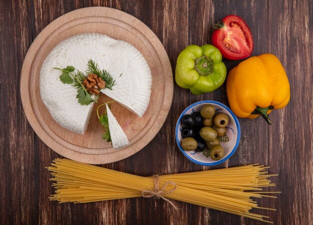 Top view feta cheese on a stand with bell peppers  tomatoes  olives and raw spaghetti on a wooden background