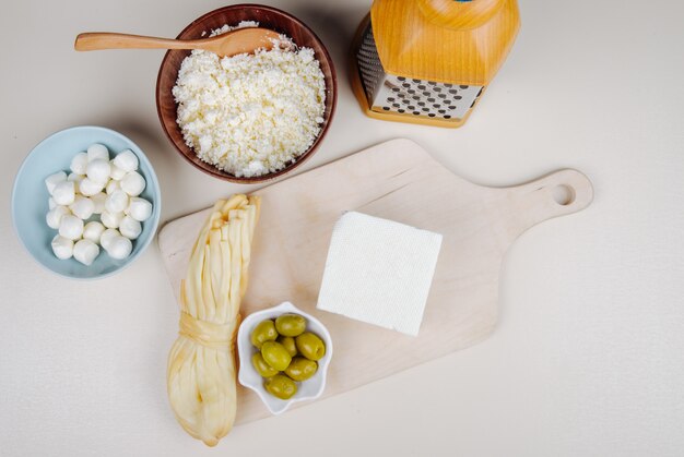 Top view of feta cheese with pickled olives on a wooden cutting board and string cheese , cottage cheese in a bowl and mozzarella with grater on white table