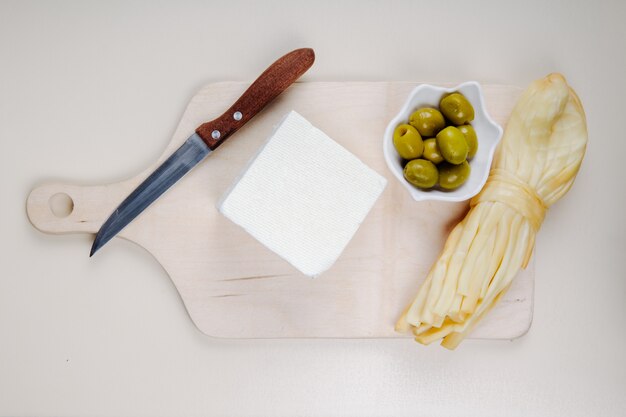 白いテーブルに木製のまな板にストリングチーズ、ピクルスオリーブ、包丁とフェタチーズのトップビュー