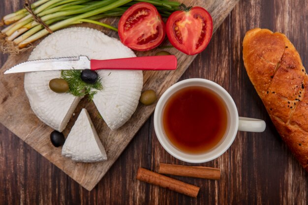 Вид сверху сыр фета с помидорами, оливками и зеленым луком на подставке с чашкой чая и буханкой хлеба на деревянном фоне