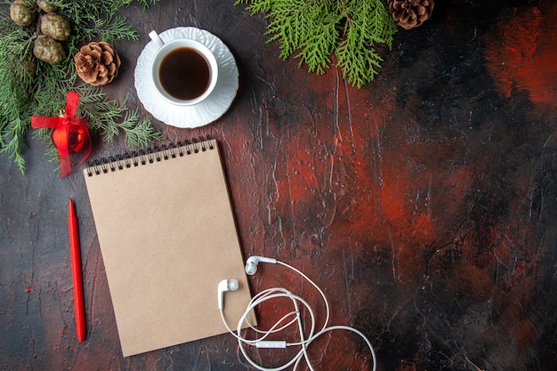Top view of fir branches a cup of black tea decoration accessories white headphone and gift next to notebook with pen on dark background