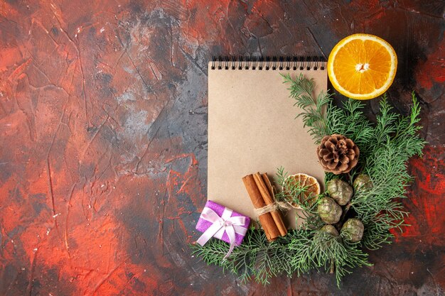 Top view of fir branches purple color gift and closed spiral notebook cinnamon limes and cut orange on the left side on red background