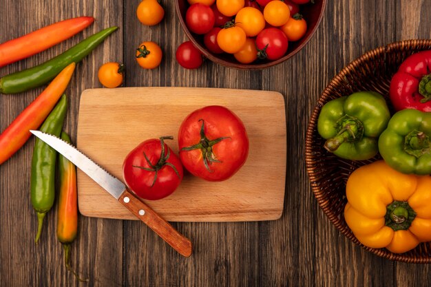 Top view of flavored red large sized tomatoes on a wooden kitchen board with knife with cherry tomatoes on a wooden bowl and bell peppers on a bucket on a wooden wall