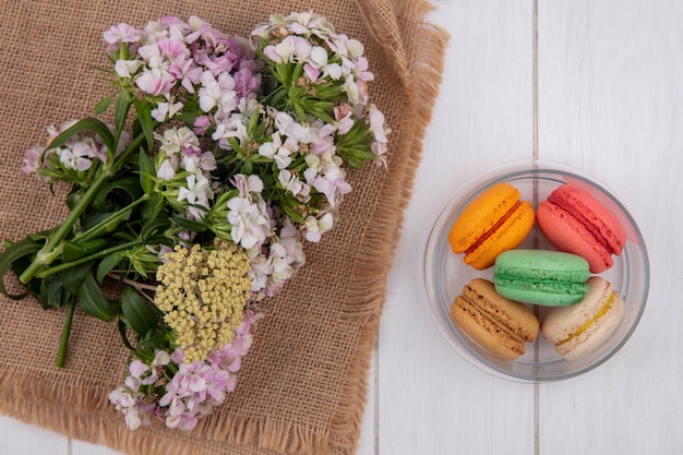 Top view of flowers on a beige napkin with colored macarons in a jar on a white surface