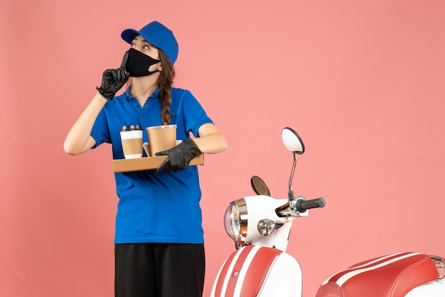 Top view of focused courier girl wearing medical mask gloves standing next to motorcycle holding coffee small cakes on pastel peach color background