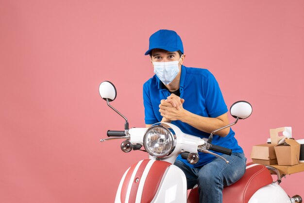 Top view of focused courier man in medical mask wearing hat sitting on scooter on pastel peach background
