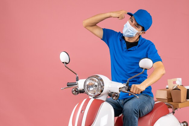 Top view of focused delivery guy in medical mask wearing hat sitting on scooter on pastel peach background