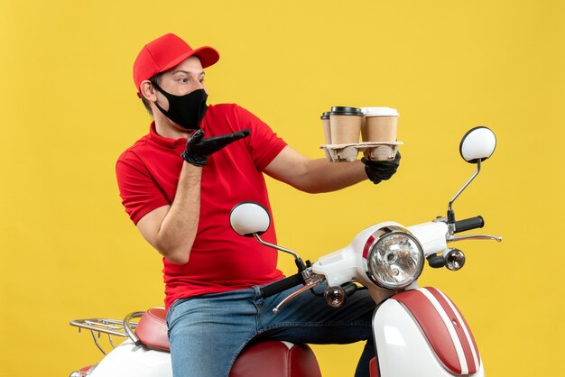 Top view of focused delivery guy wearing uniform and hat gloves in medical mask sitting on scooter showing orders