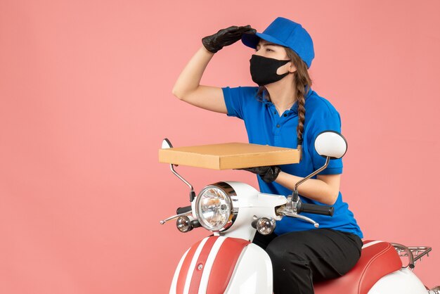 Top view of focused female courier wearing medical mask and gloves sitting on scooter delivering orders on pastel peach background