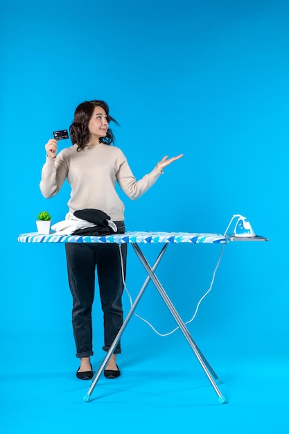 Top view of focused young woman standing behind the ironing board and showing bank card pointing something on the left side on blue wave background