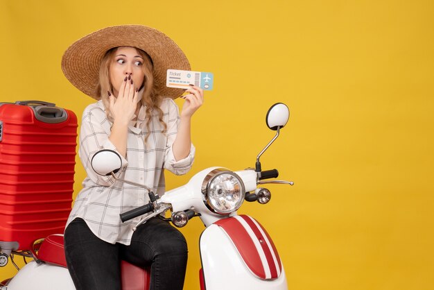 Top view of focused young woman wearing hat and sitting on motorcycle and holding ticket on yellow 