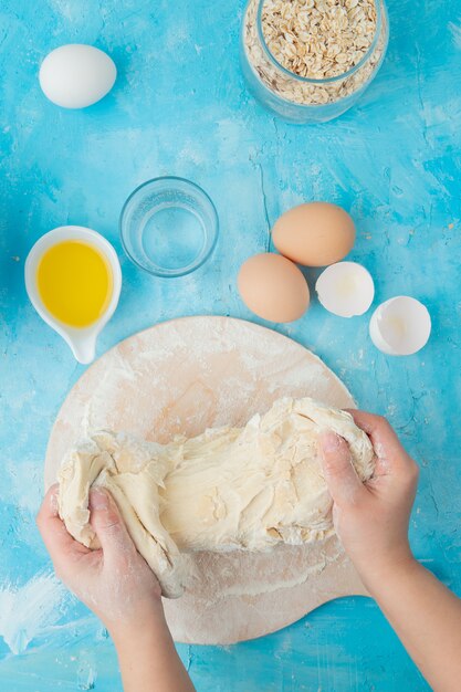 Взгляд сверху продуктов как овсяные хлопья яичка масла с тестом руки женщины замешивая на голубой предпосылке