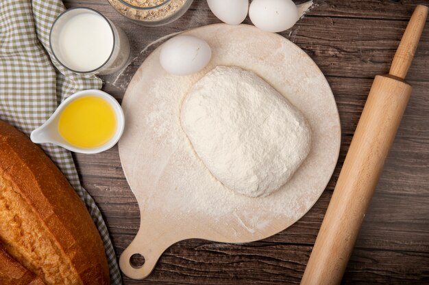 Top view of foods as milk butter baguette bread egg with dough on cutting board and rolling pin on wooden background