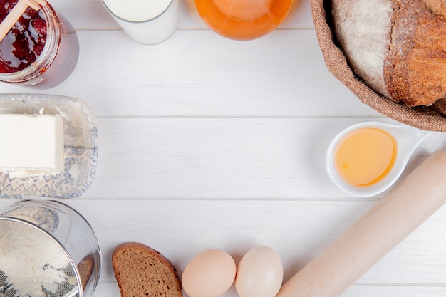 Top view of foods as strawberry jam milk butter flour cob and rye breads eggs and rolling pin on wooden background with copy space