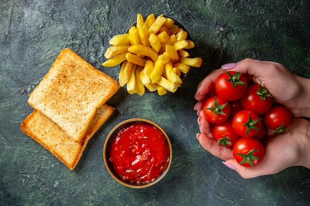 Top view french fries with toasts and red cherry tomatoes in female hands on dark surface