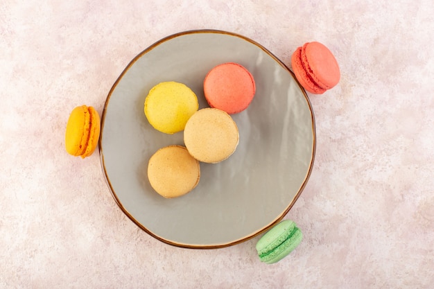 Вид сверху французские макароны внутри круглой тарелки на розовом столе, сладком бисквитном сахаре