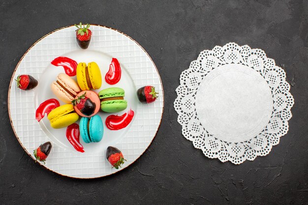 Top view French macaroon chocolate-covered strawberries and French macaroons next to the lace doily on the dark table