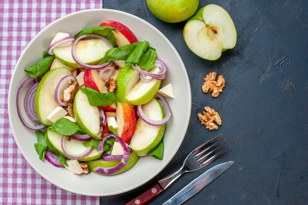 Салат из свежих яблок на круглой тарелке, вид сверху, зеленые яблоки, грецкий орех, фиолетовая и белая клетчатая скатерть, вилка и нож на темном столе