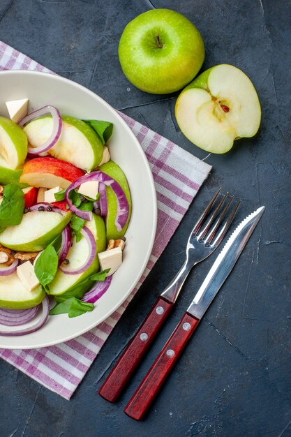 둥근 접시에 있는 신선한 사과 샐러드 보라색과 흰색 체크 무늬 식탁보 녹색 사과 포크와 나이프는 어두운 탁자에 있습니다.