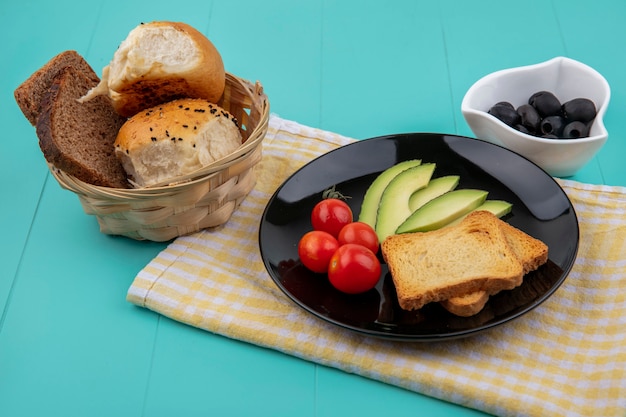 Top view of fresh avocado slices with tomatoes and toasted bread slices on black plate on yellow checked tablecloth with black olives on white bowl on blue