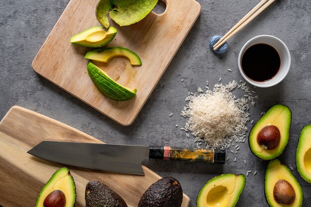 Top view of fresh avocados, a cutting board and knife, rice, and chopsticks on a grey surface