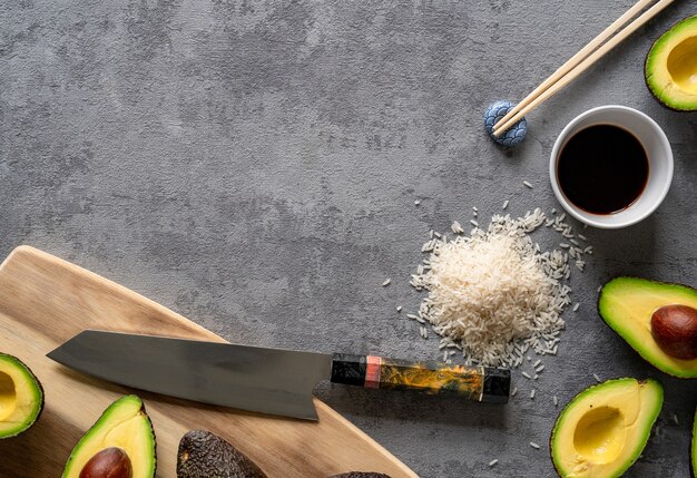 Top view of fresh avocados, a cutting board and knife, rice, and chopsticks on a grey surface