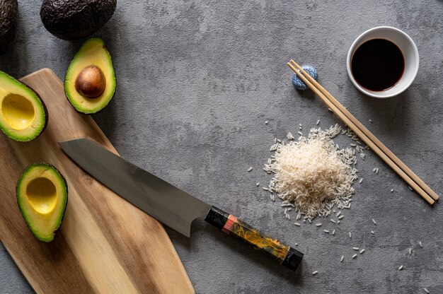 Top view of fresh avocados, a cutting board and knife, rice, and chopsticks on a grey surface