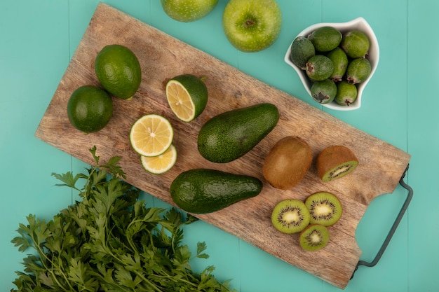 Top view of fresh avocados with limes and kiwis on a wooden kitchen board with feijoas on a bowl with apples and parsley isolated on a blue wall